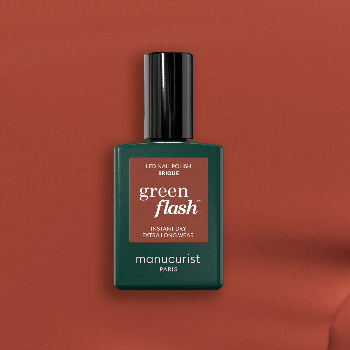 A bottle of Manucurist nail polish in the shade Brique is shown, featuring a dark green rectangular bottle with a black cap set against a brick red background.