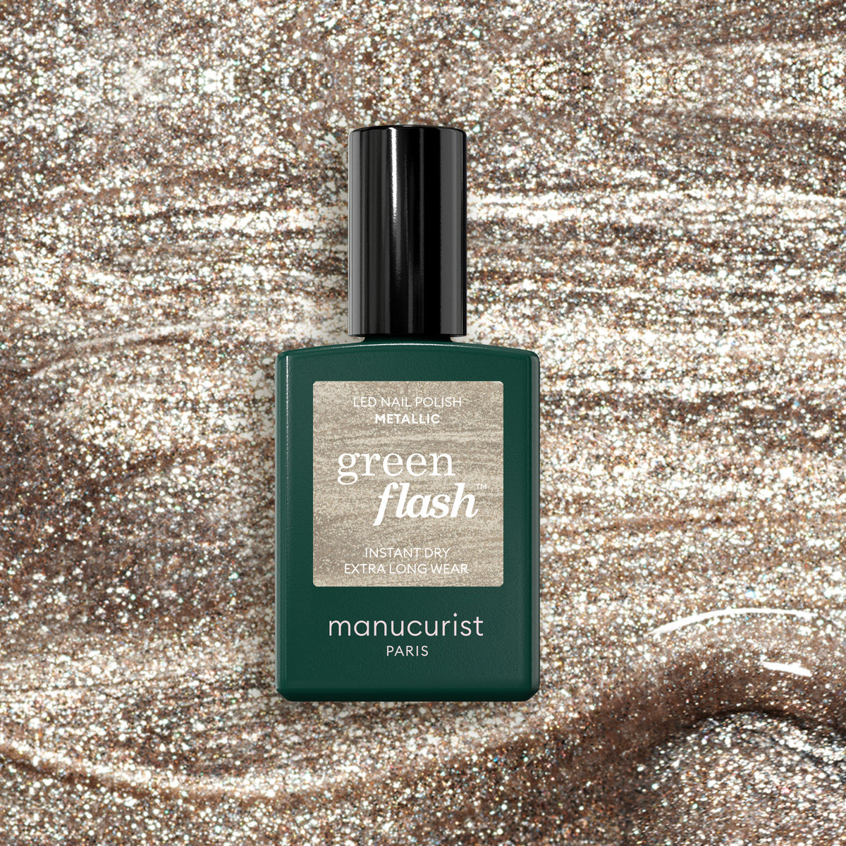 A bottle of Manucurist Metallic nail polish sits on a sparkling, shimmery metallic surface with cool gold highlights matching the polish&