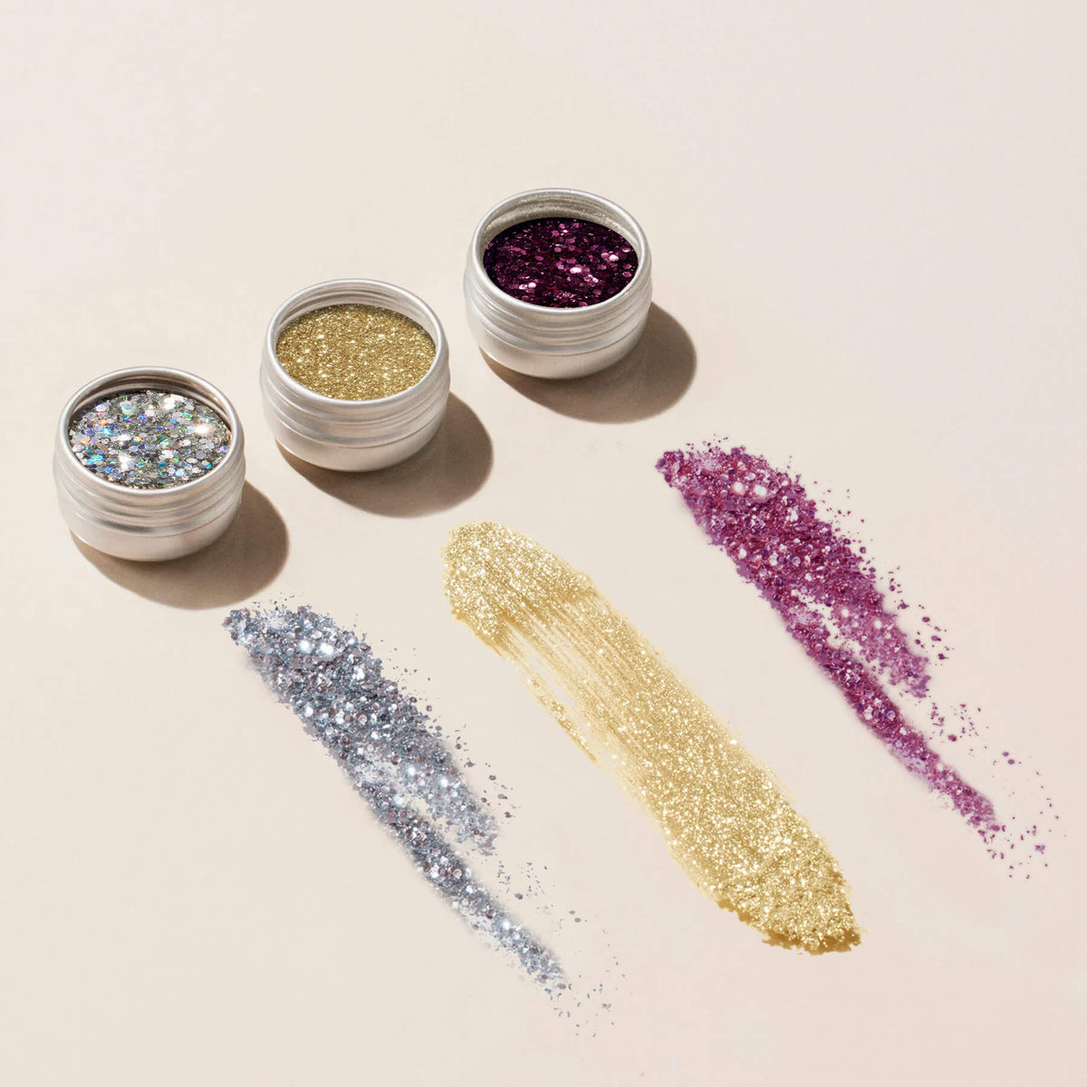 Three small jars from the Manucurist Glitters and Effect Powders Trios—silver, gold, and purple—are open in a row, each with matching powder streaked on the surface below.