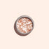 A round container of Manucurist Blush biodegradable glitter features coarse pink and white crystals with eco-friendly sparkle accents, resembling bath salts or body scrub, set against a light beige background.