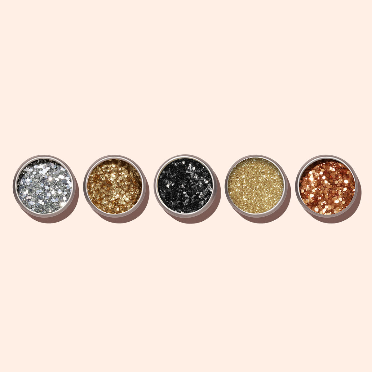 Five small round Manucurist Gold Biodegradable Glitter containers for nail art, in gold, black, yellow-gold, silver, and copper shades, are arranged in a row on a light beige background.