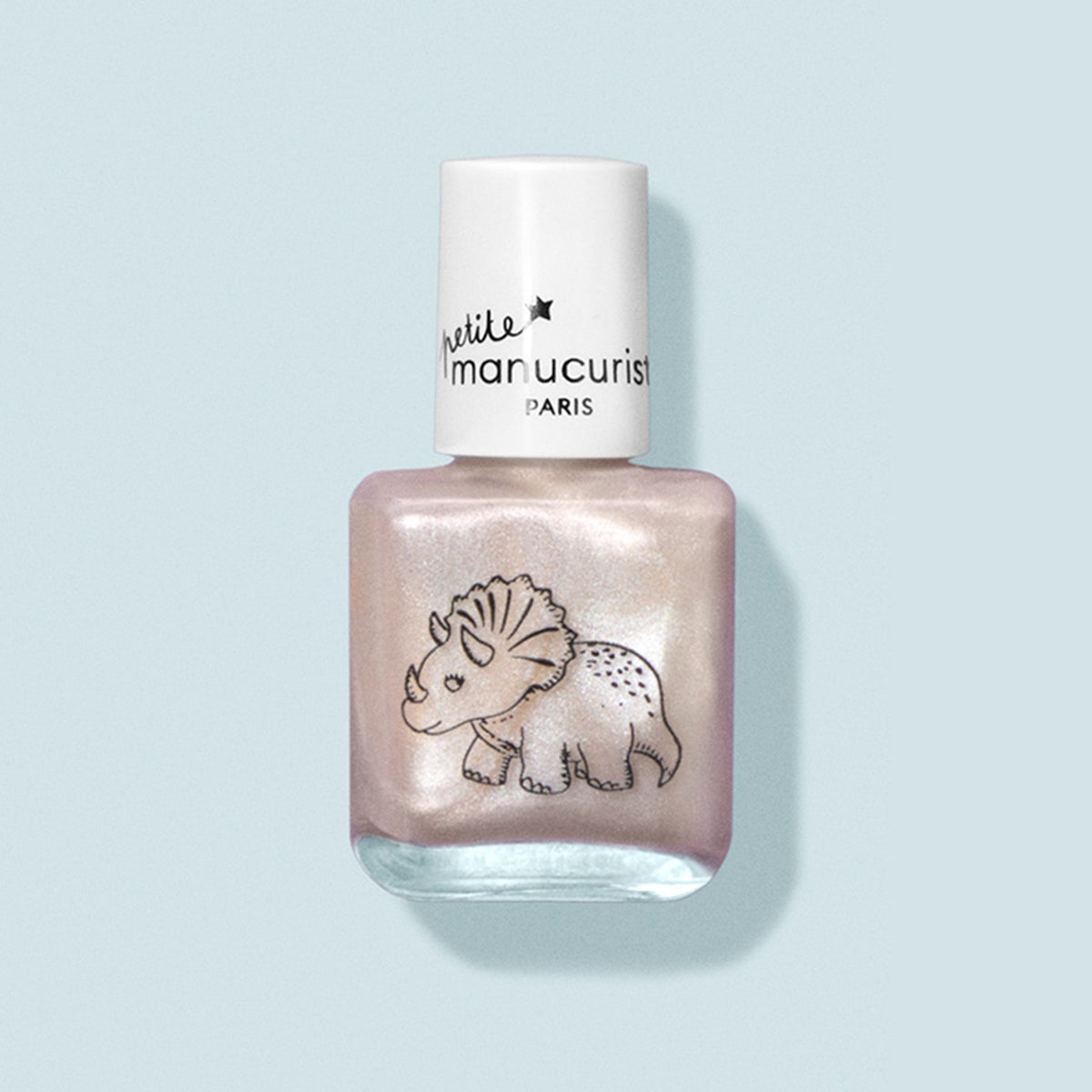A bottle of Manucurist Pénélope the Triceratops kids nail polish in pale pink, featuring a cute triceratops design and white cap, made with natural ingredients. Shown against a light blue background.