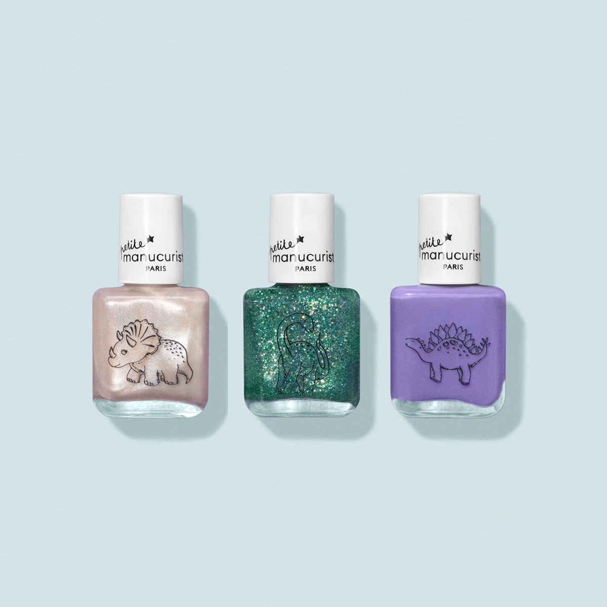 Three bottles of Manucurist’s Wild Life Collection kids nail polish, each with a white cap and dinosaur illustration, in pearly white, glittery green, and lavender purple shades—all set against a light blue background.