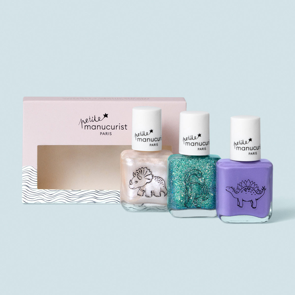 Three bottles from the Manucurist Wild Life Collection, featuring dinosaur designs, sit next to a pink and white windowed box against a light blue background—a gentle, washable nail polish set perfect for kids.