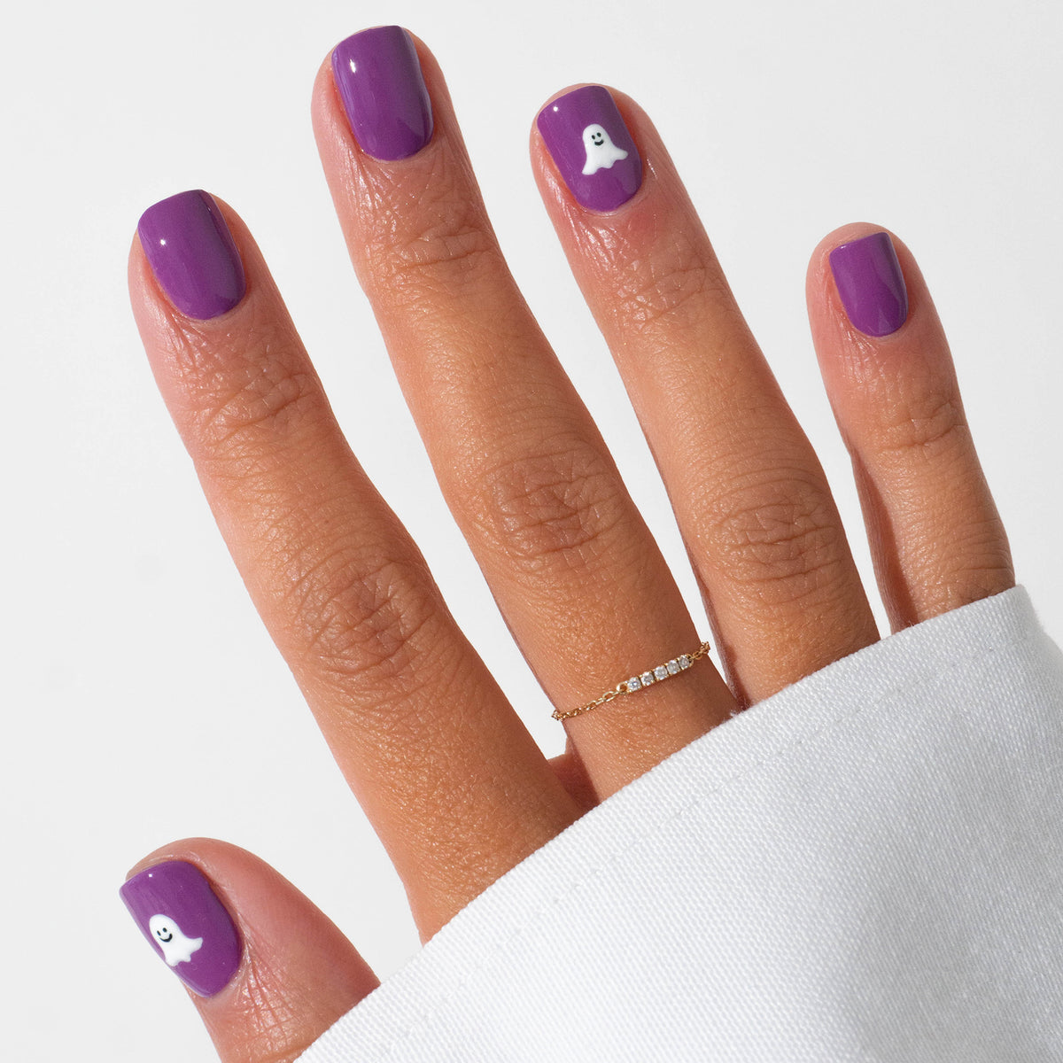 A hand displays Halloween nail art with Manucurist’s Witchy  Wicked Set—two purple gel-polished nails feature white ghost designs. A dainty gold ring adorns the hand, partly covered by a white sleeve.