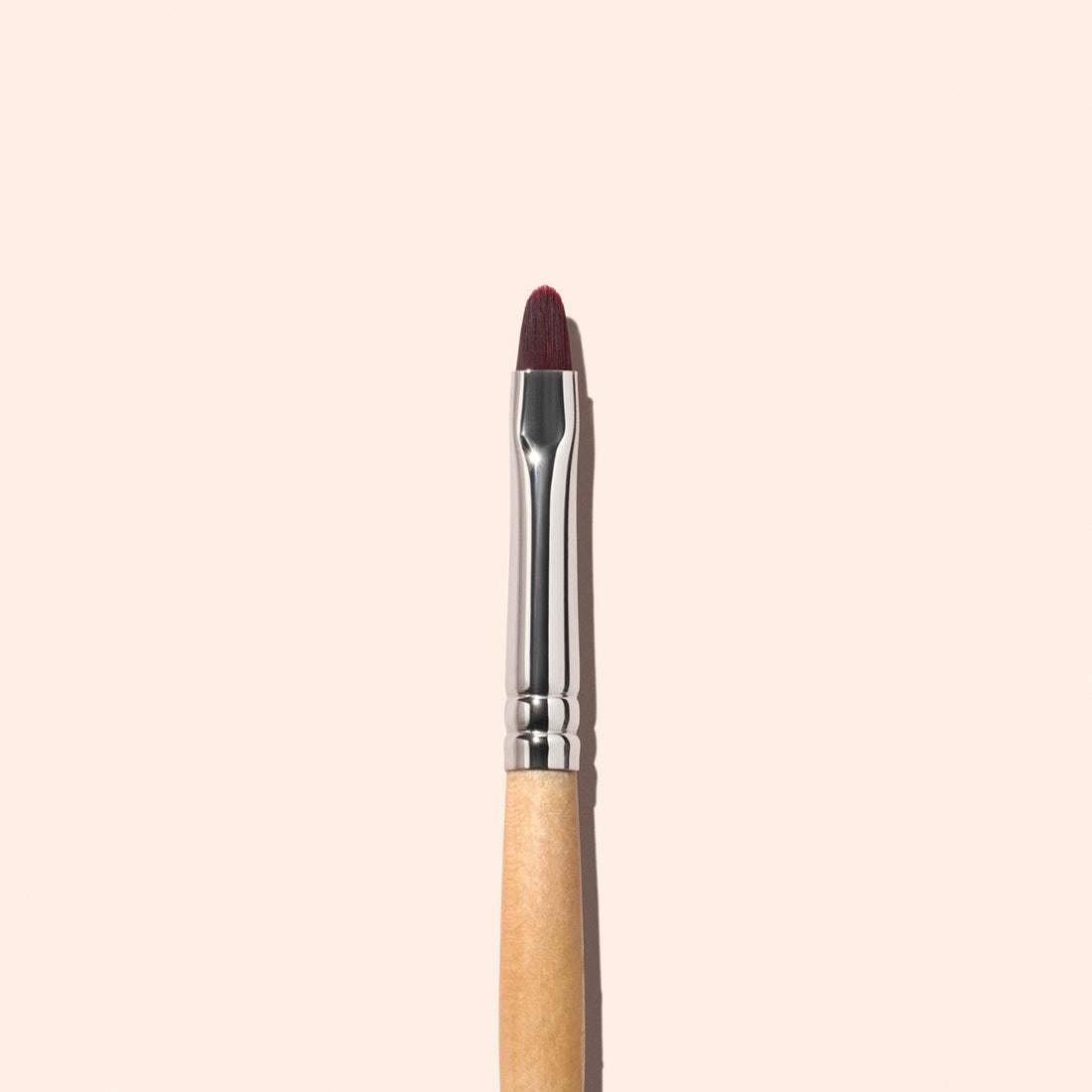 A Manucurist Flat Brush featuring a wooden handle, silver ferrule, and a small, rounded dark bristle tip—perfect for precision tasks like manicure touch-ups or cuticle care—is positioned vertically against a light beige background.