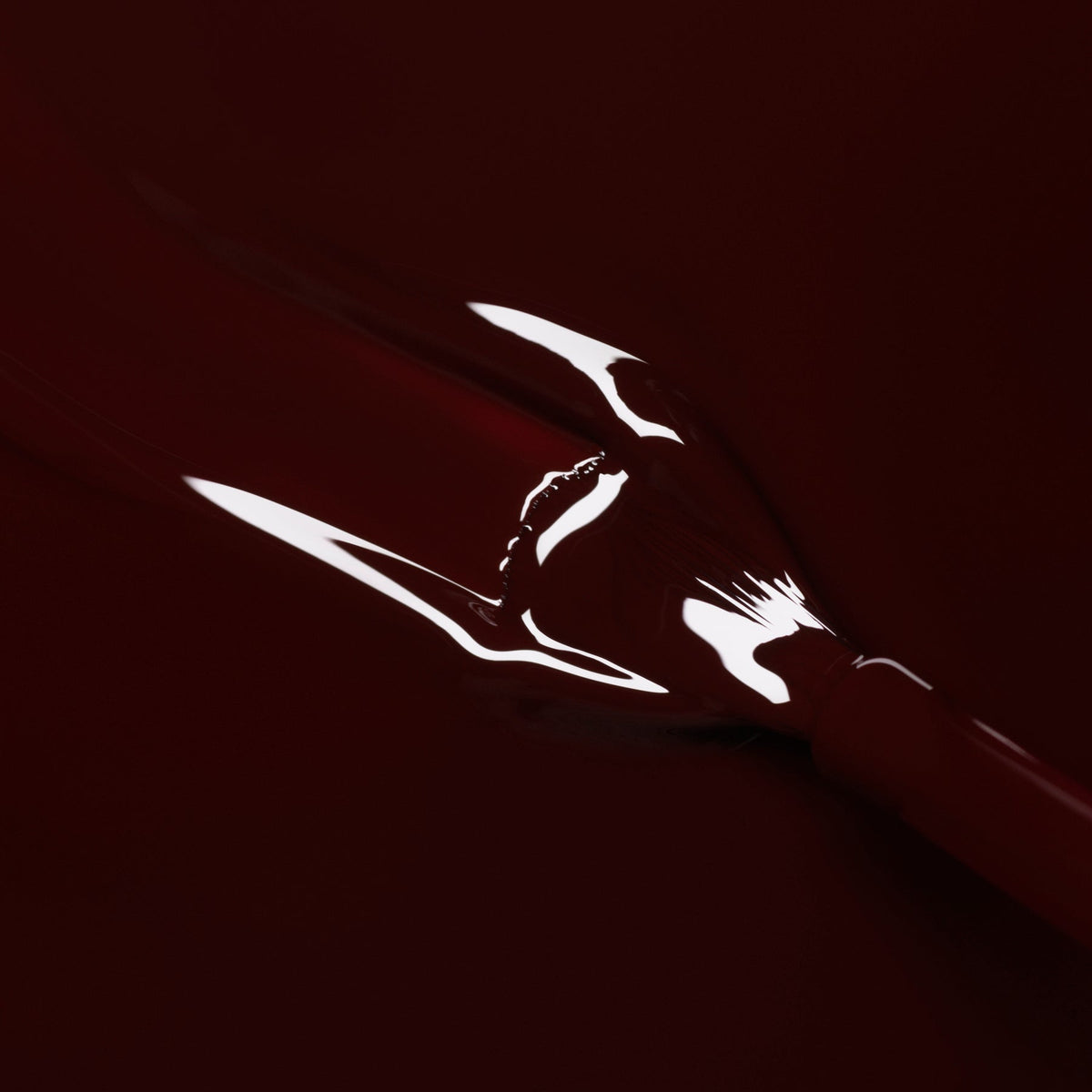 A close-up of Manucurist’s Hollyhock, a glossy deep red nail polish, spread on a dark surface with a reflection in the liquid that resembles the outline of lips.