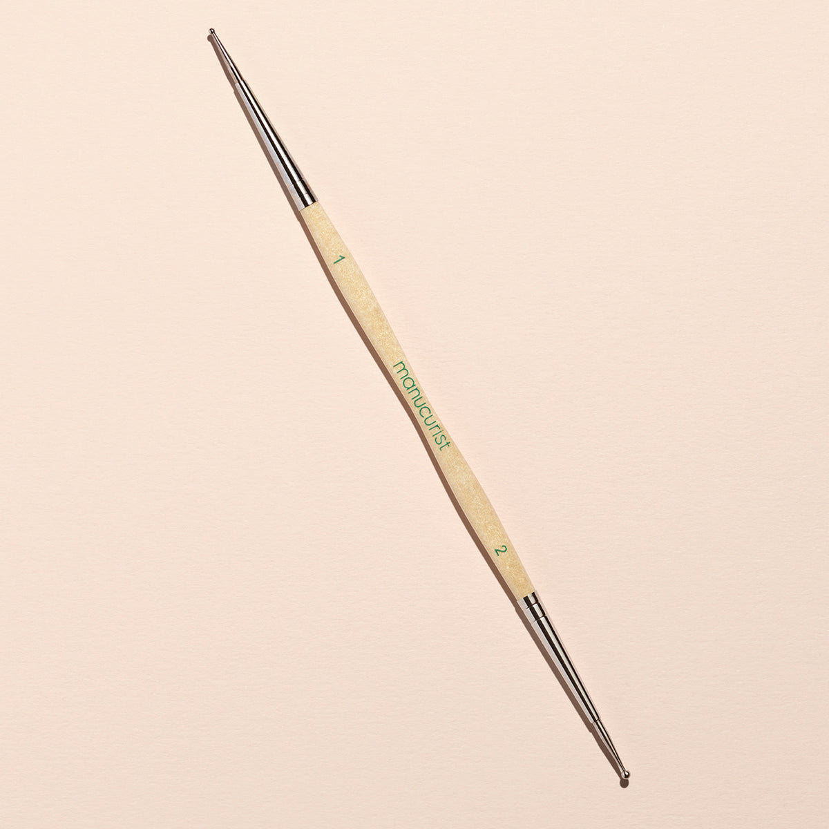 The Manucurist Dotting Tool features a wooden handle and double-ended metal tips in sizes 1 and 2. Placed diagonally on a plain beige background, it’s ideal for creating precise nail art designs.