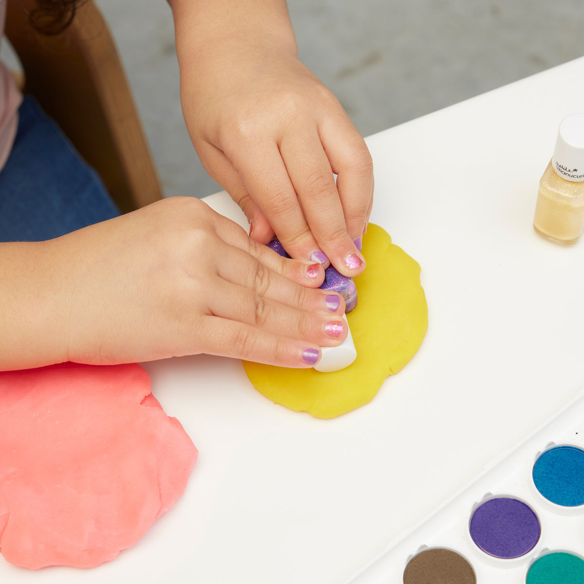A child with glittery nails uses the Manucurist Betty Scarlett Stella set, pressing a white stamp into yellow playdough. Pink playdough, watercolor paints, and shimmery powder nearby add extra sparkle to this creative scene.