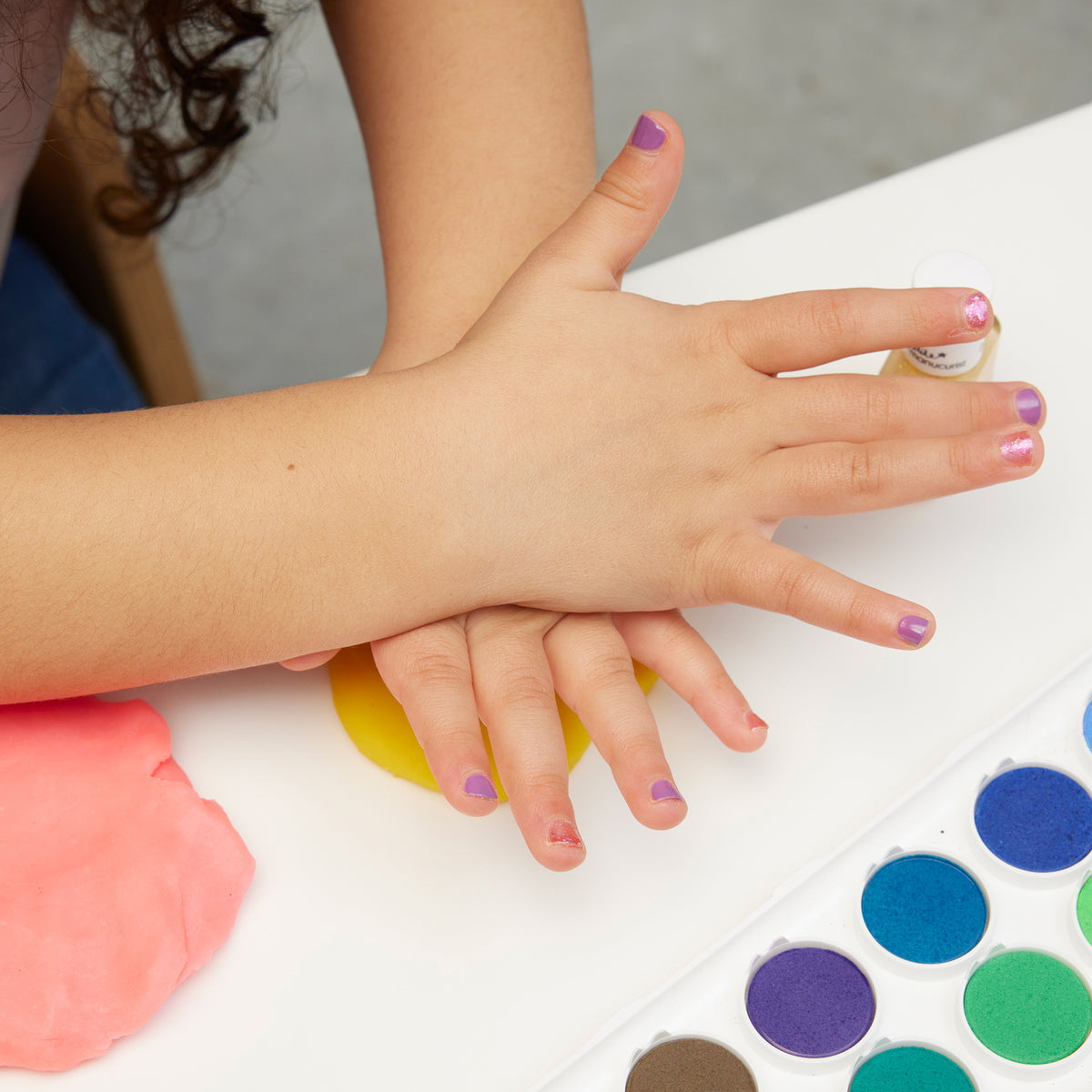A child with glittery nails from the Manucurist Bonnie Stella Janis set presses their hands onto a yellow playdough ball at a table with paint pots and pink playdough nearby.
