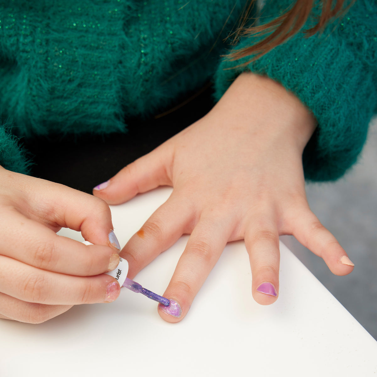 A child in a green sweater paints their nails with glitter polish from the Manucurist Bonnie Stella Janis set at a white table.