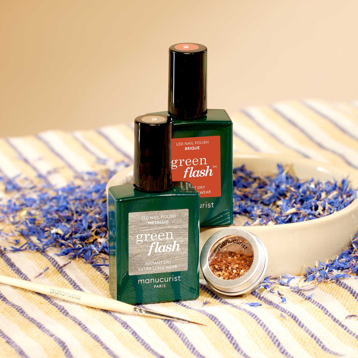 Two bottles of Manucurist Oriental Vibe nail polish from the Autumn 23 collection and a small open jar of biodegradable glitter rest in a bowl with dried blue petals on a striped cloth. A manicure tool is placed nearby.