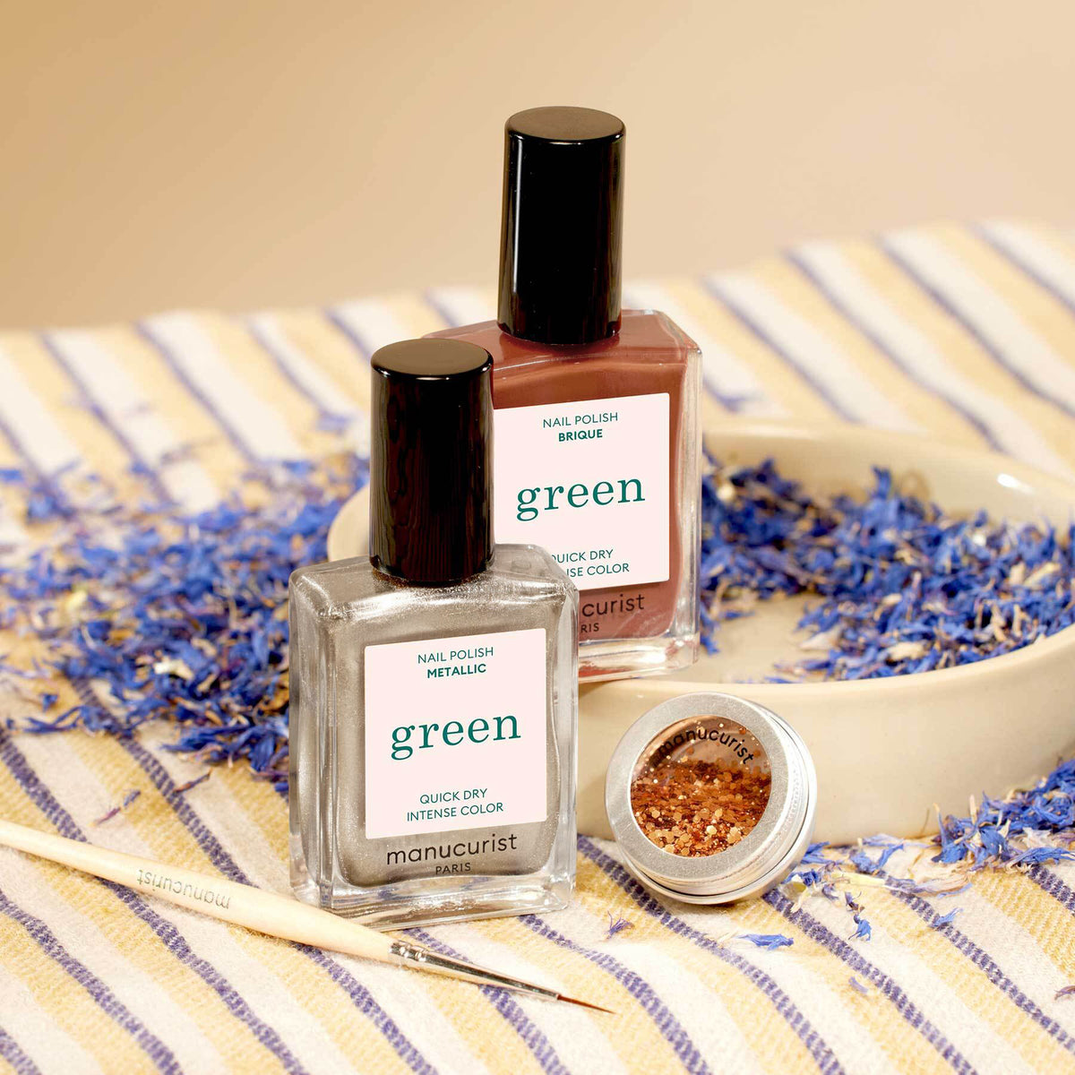 Two bottles of Manucurist’s Oriental Vibe nail polish—one metallic silver, one brick red—rest on striped fabric near an open jar of biodegradable glitter, a wooden brush, and scattered blue flower petals.