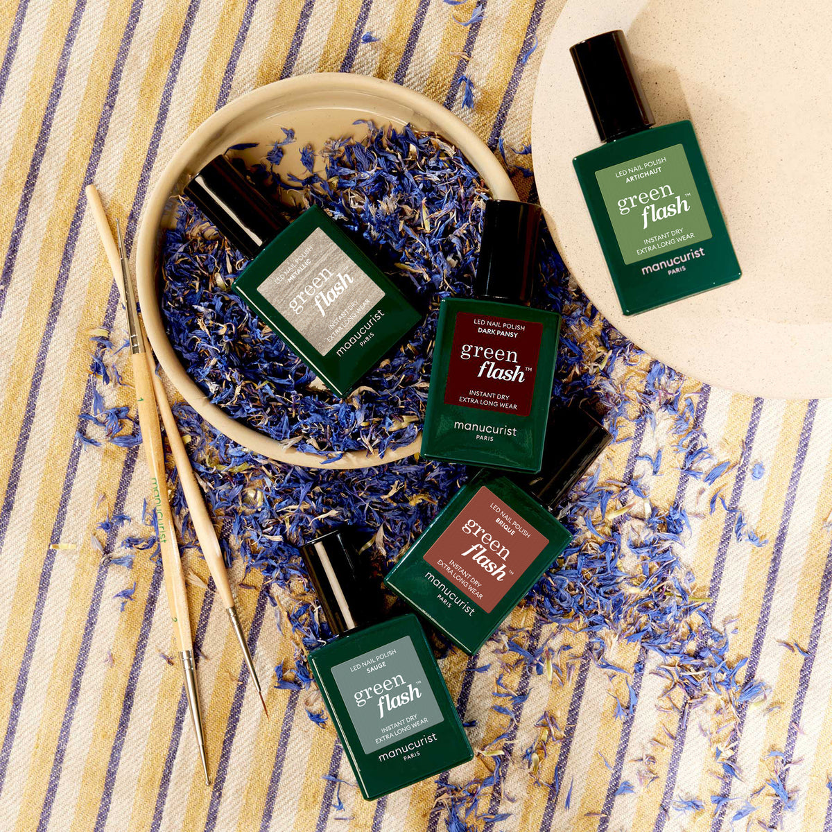 Five Manucurist Spice Lover bottles are artfully arranged on dried blue petals with chopsticks and a beige plate, set on striped fabric—a perfect scene to inspire your next nail art look.
