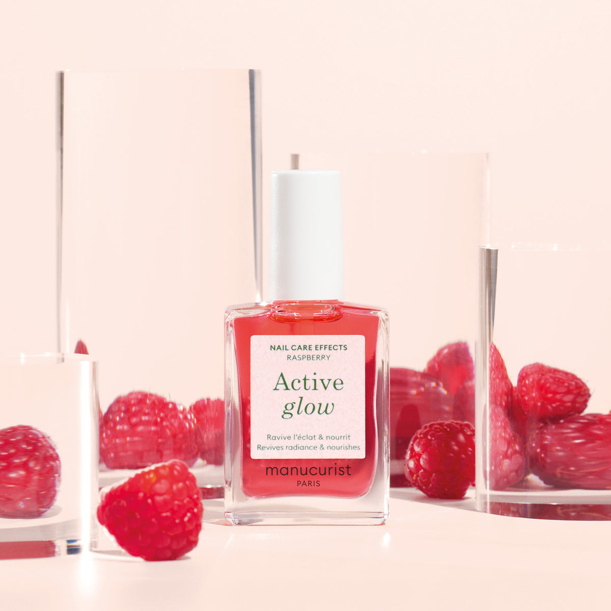 A bottle of Manucurist Active™ Glow Raspberry nail polish, a nail enhancer created to boost natural radiance, is surrounded by fresh raspberries and clear glass blocks on a light pink background.