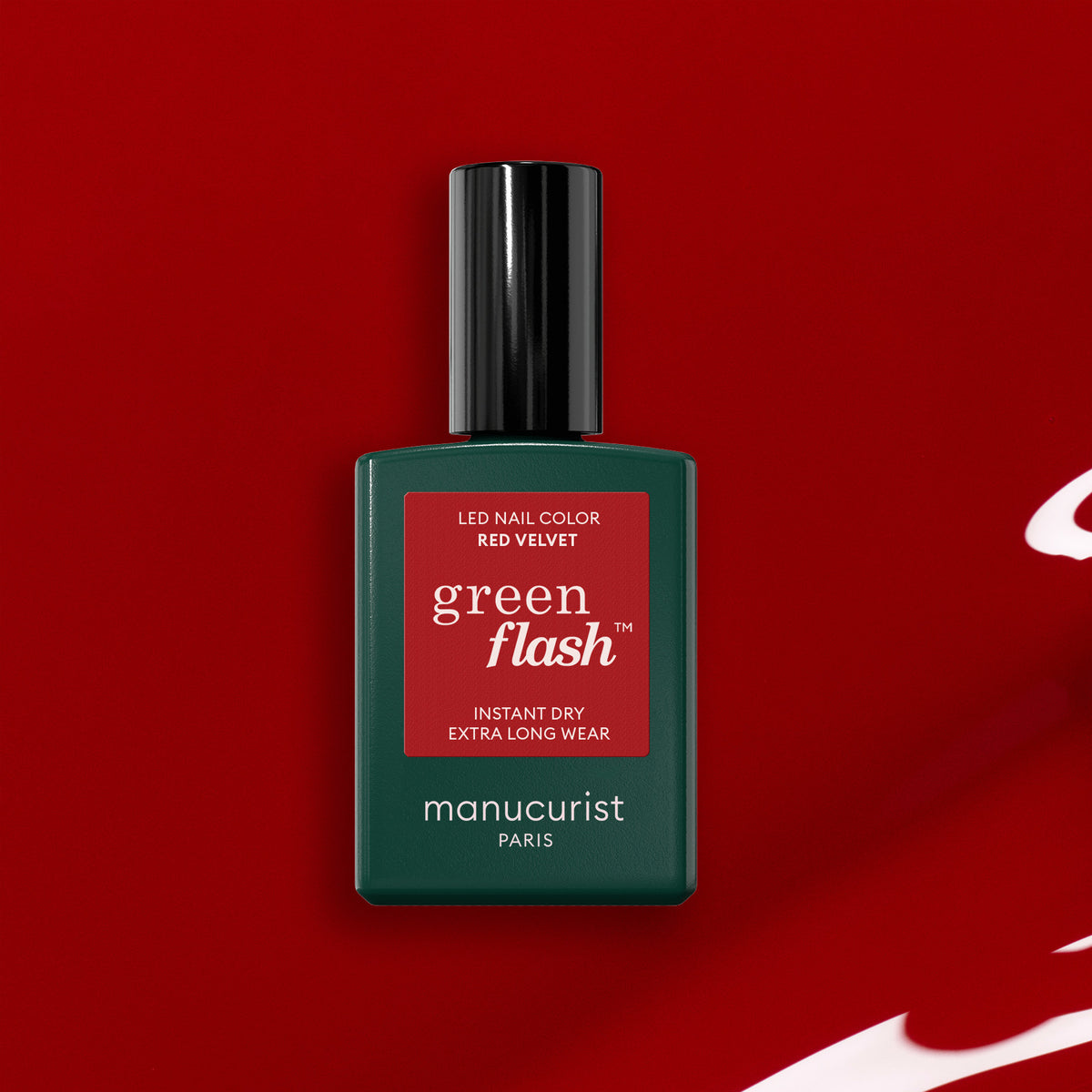 A bottle of Manucurist Red Velvet Green Flash LED gel polish, featuring a dark green label and black cap, rests on a glossy red background with a splash of velvet red gel polish.