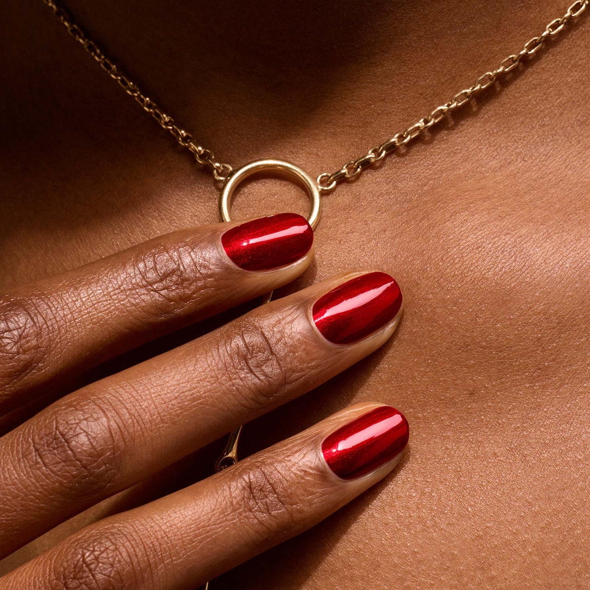 A close-up of a hand with Manucurist’s Red Hibiscus nail polish rests on a person’s chest, drawing attention to a gold chain necklace with a circular pendant.