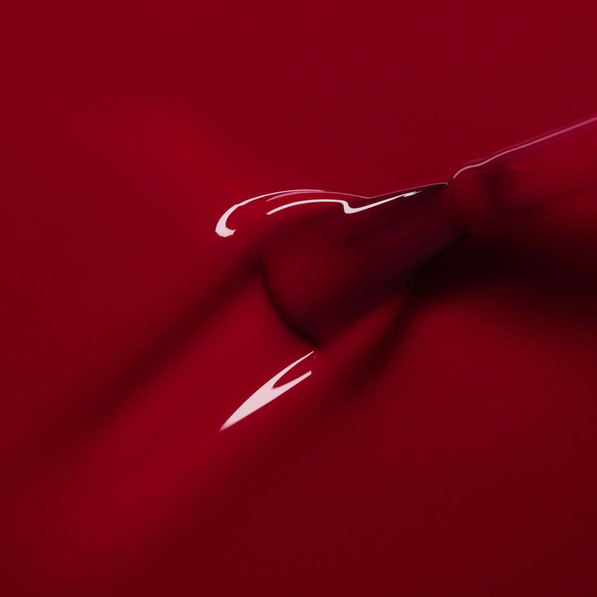 A close-up of a Manucurist Pomegranate dropper releasing glossy red liquid onto a smooth red surface, with highlights reflecting on the liquid.