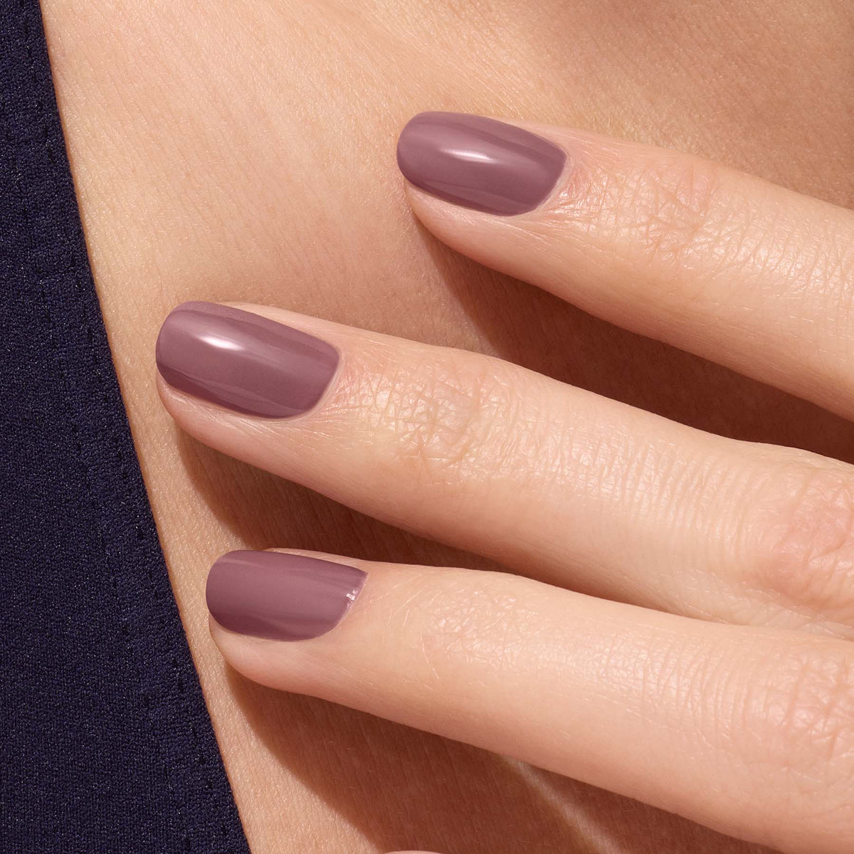 A close-up of a hand with neatly manicured nails painted in Manucurist’s Rose Mountbatten, a glossy pinky taupe shade, gently resting against bare skin and a dark fabric background.