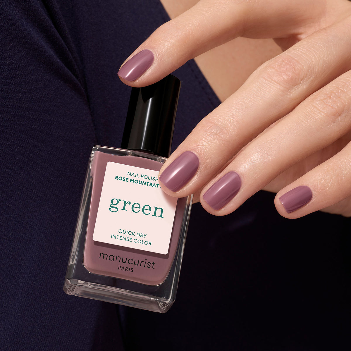 A hand with short, neat nails painted in Manucurist Rose Mountbatten holds a matching bottle. The person wears a dark purple top.