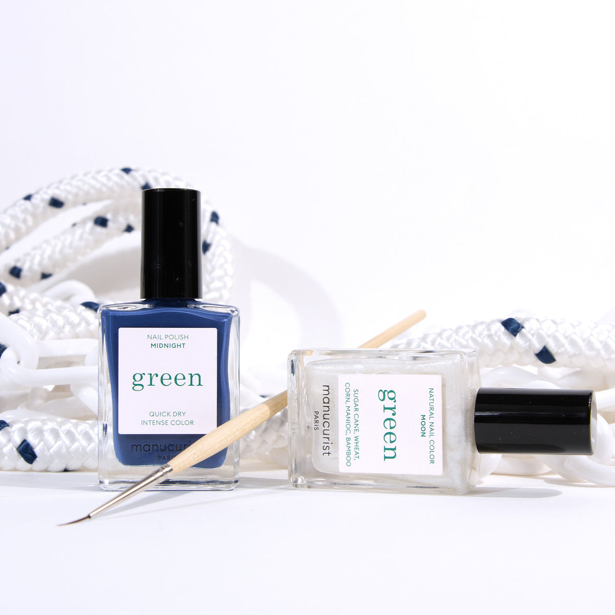 Two bottles of Manucurist’s Midnight Swim nail polish—one blue, one clear—rest on a white surface next to a Nail Art Liner Brush, with a coiled rope featuring blue accents visible in the background.