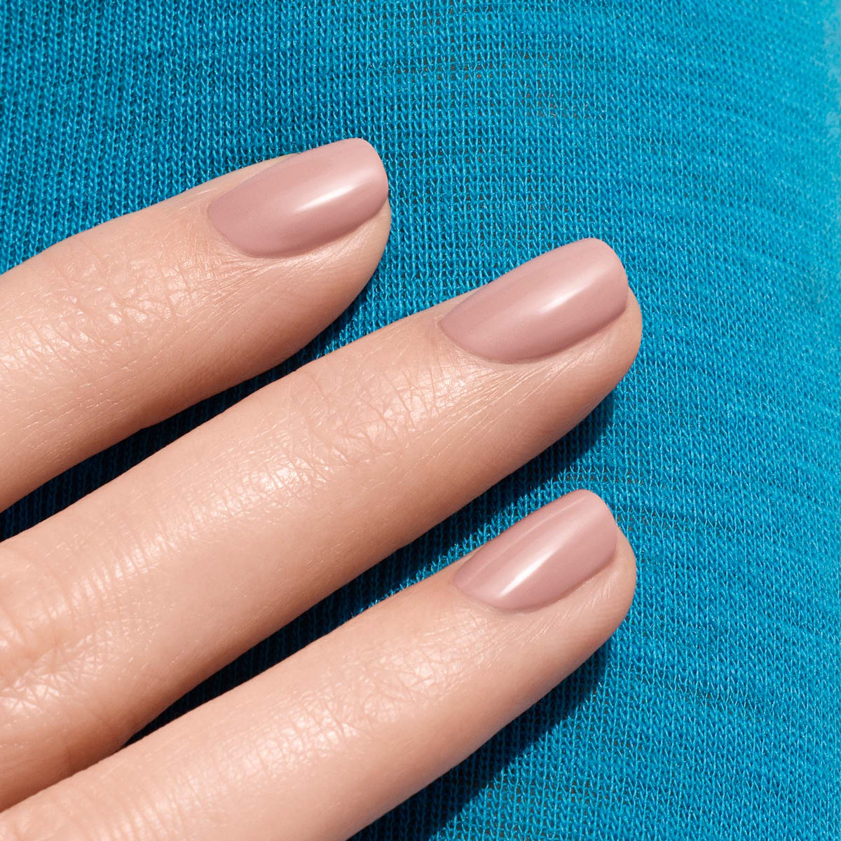 Three fingers with short, neatly manicured nails painted in Manucurist’s Shell Beige, a delicate pinkish beige shade, rest on textured blue fabric.