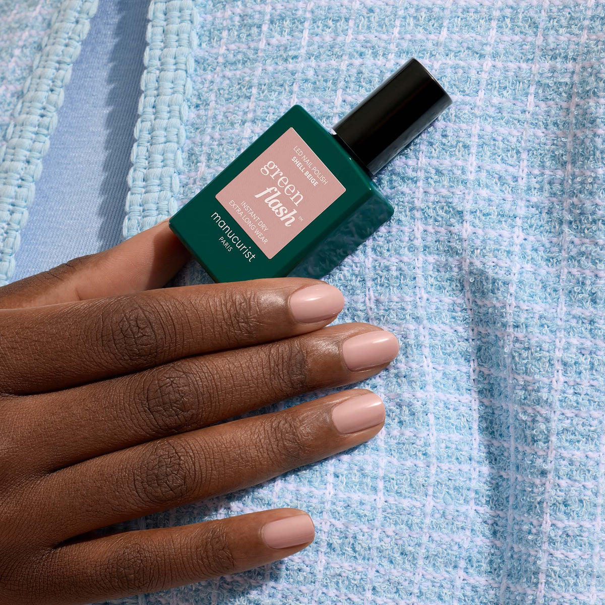 A hand with neatly manicured nude nails holds a green bottle of Manucurist Green Flash gel polish in the shade Shell Beige, set against a light blue textured fabric background.