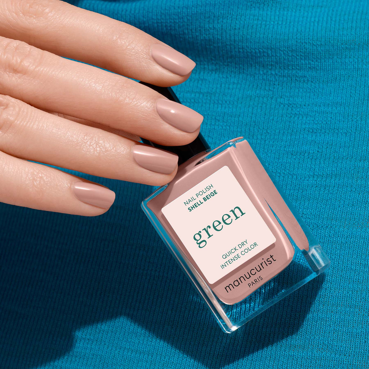 A hand with neatly manicured nails painted in a delicate pinkish beige holds a bottle of Manucurist Shell Beige nail polish, set against a blue fabric background.