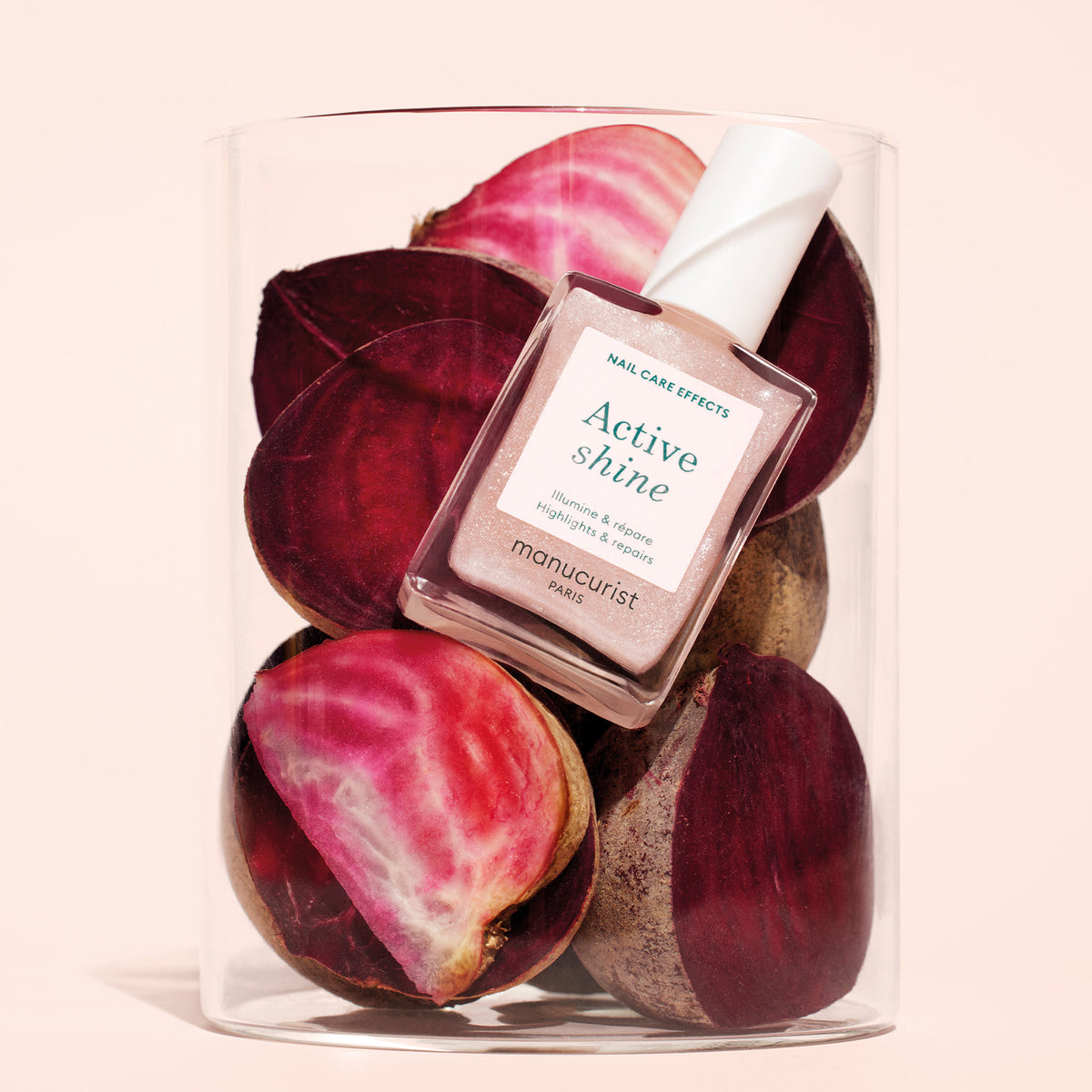 A bottle of Manucurist Active™ Shine Opal plant-based nail polish rests on sliced beets inside a clear glass container against a pale background.