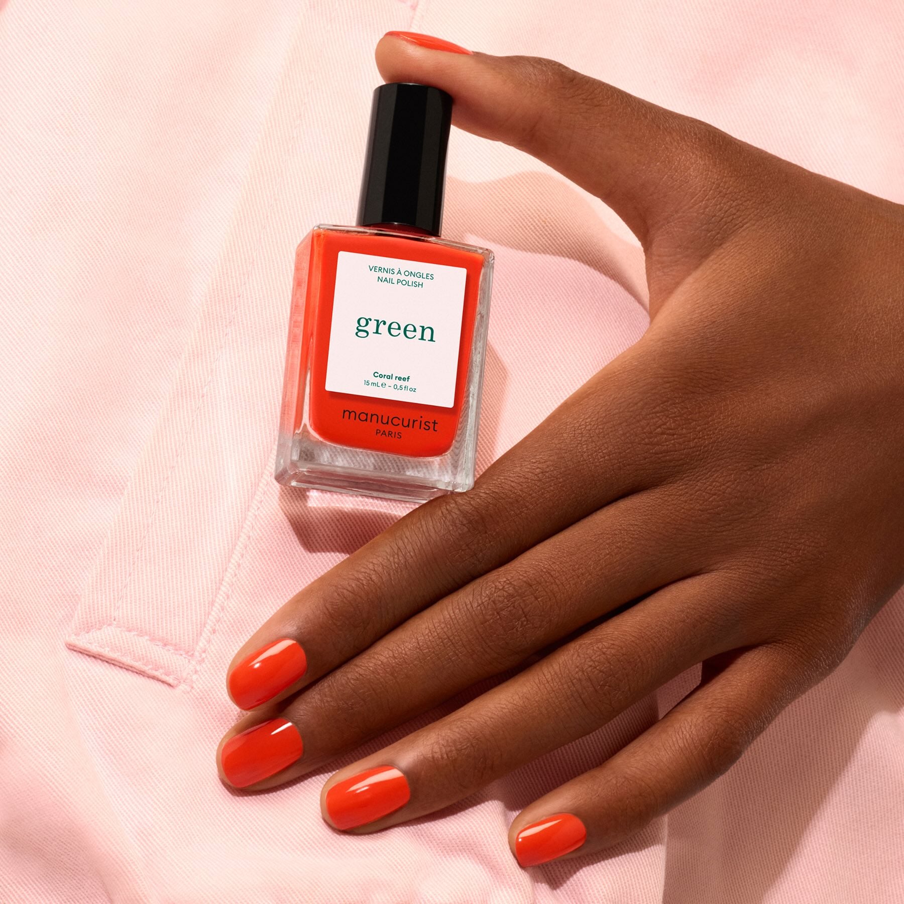 A hand with neatly manicured nails painted in vibrant coral holds a matching bottle of Manucurist Coral Reef nail polish against a light pink fabric background.