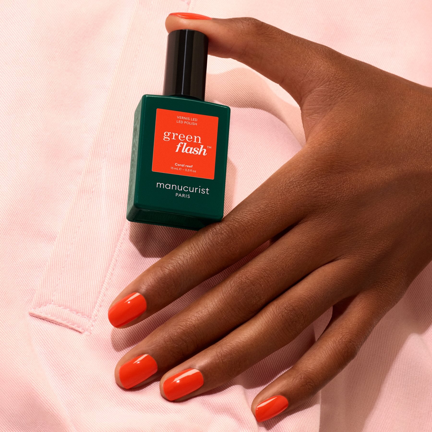 A hand with neatly manicured, bright orange nails holds a bottle of Manucurist Coral Reef polish against a light pink fabric background.