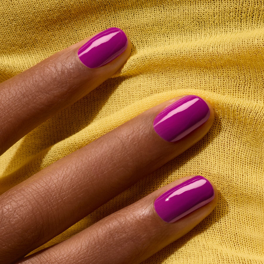 A close-up of a hand with neatly manicured nails painted in "Fizz" by Manucurist, a violet gel polish with magenta undertones, resting on soft yellow fabric.