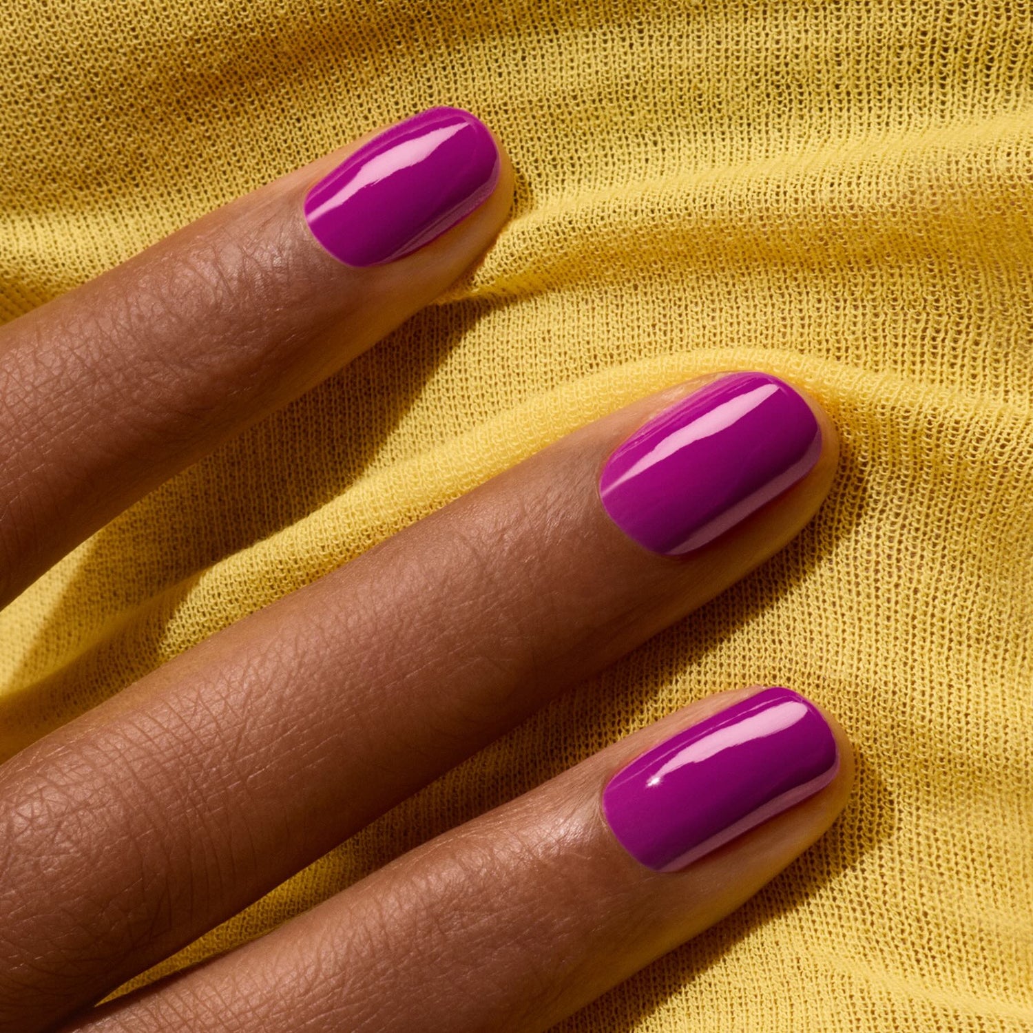 A close-up of a hand with neatly manicured nails painted in "Fizz" by Manucurist, a violet gel polish with magenta undertones, resting on soft yellow fabric.