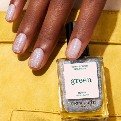 A hand with glittery pastel nails and sheer accents holds a bottle of Manucurist Mermaid nail polish against a yellow fabric backdrop.