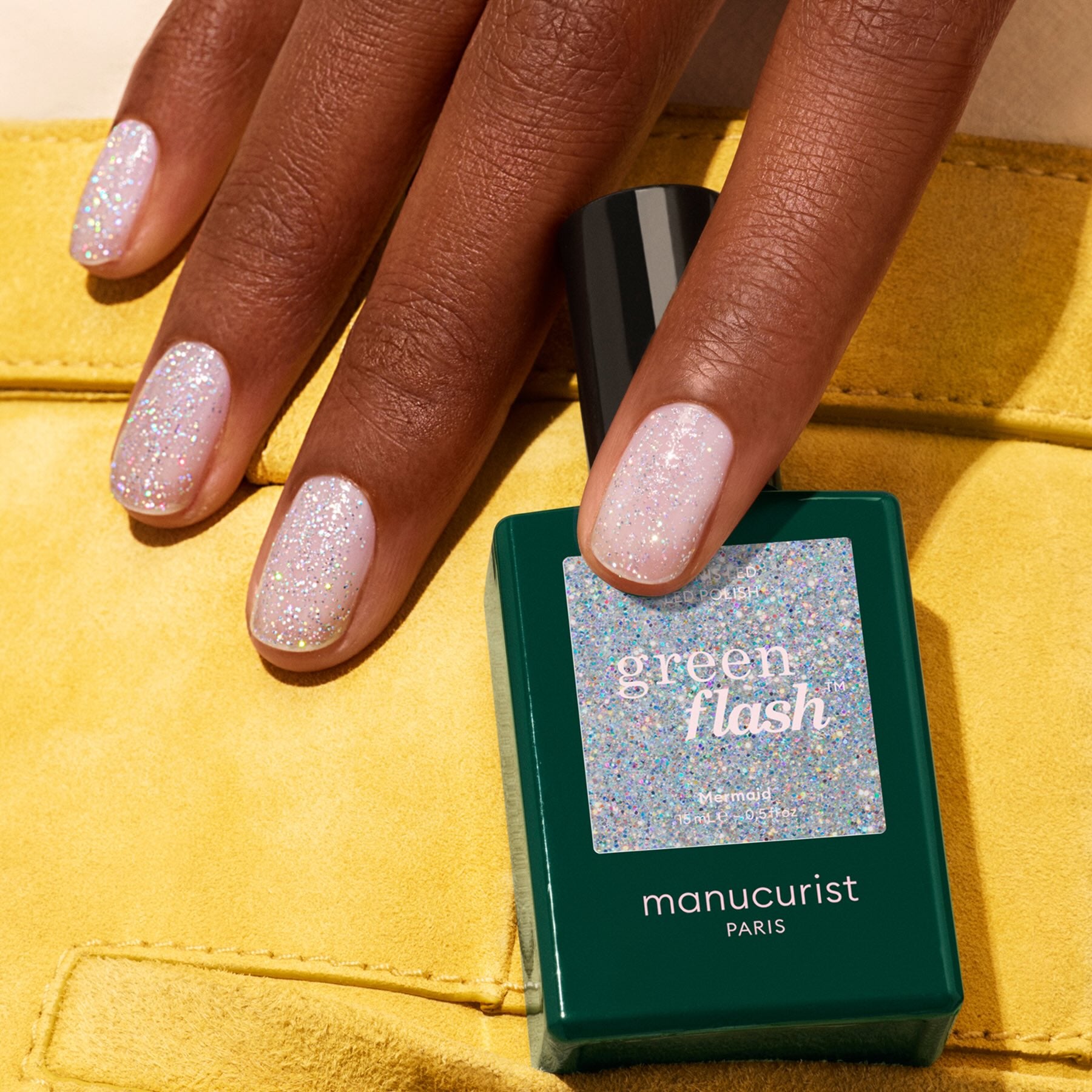 A hand with short, manicured nails painted a sparkly light shade rests by a green Manucurist bottle labeled "Mermaid" on yellow fabric.