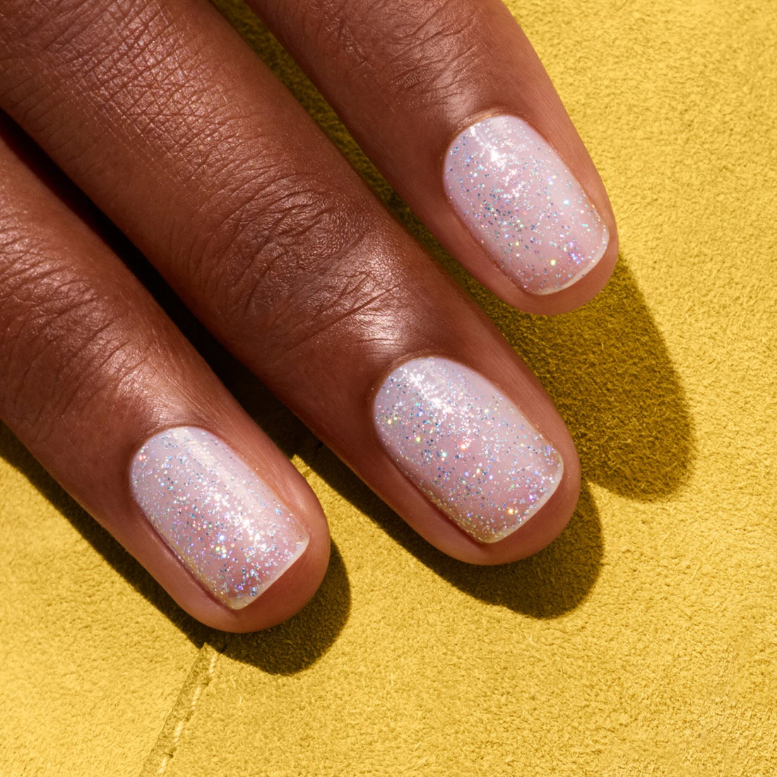 Three fingers with short, rounded nails painted in Manucurist’s Mermaid—a pale pink sheer nail polish with glitter—against a yellow textured background.