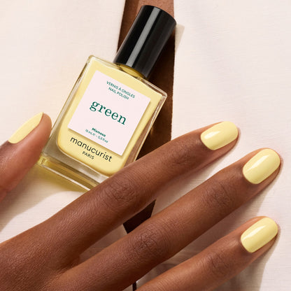 A hand with neatly manicured nails painted in Mimosa by Manucurist rests next to a bottle of the polish on light-colored fabric.