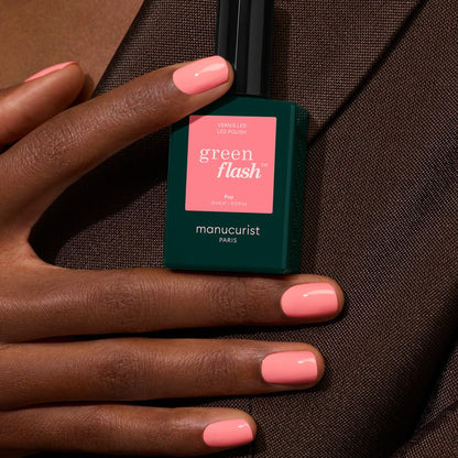 A hand with short, manicured nails painted in Pop by Manucurist holds the bottle against a brown fabric background.