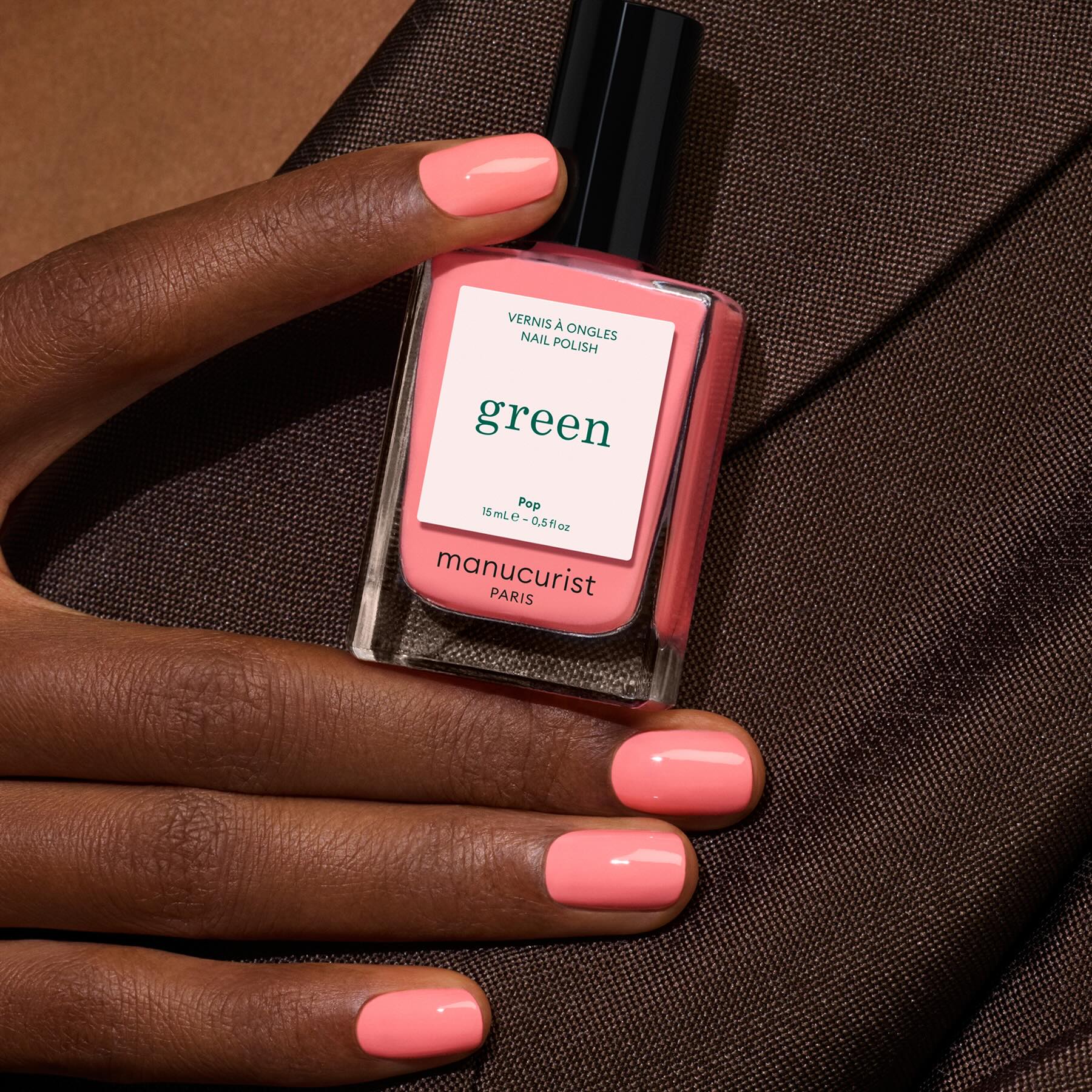 A hand with neatly manicured nails painted in vibrant coral pink holds a bottle of Manucurist nail polish in the shade Pop, with a brown fabric background.