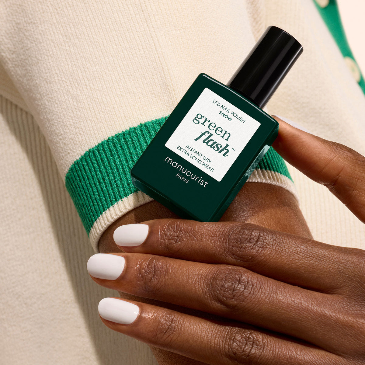 A hand with neatly manicured Snow by Manucurist pure white nails holds a dark green bottle of gel polish, set against an off-white sweater with green trim.