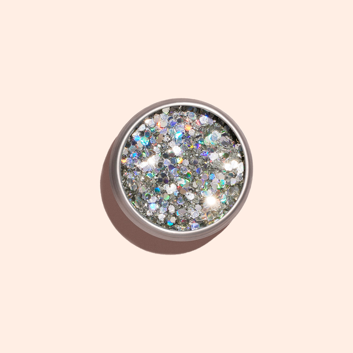 A small round container of Manucurist Holo biodegradable glitter, featuring silver and iridescent chunky sparkles, casts a soft shadow on a pale beige background.