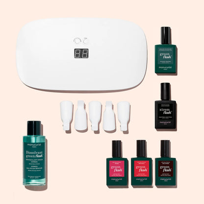 Introducing the Manucurist Starter Kit: a complete gel polish set that includes a white LED lamp, nail polish remover, four bottles of Green Flash nail polish, a vitaminized base coat for healthy nails, one Black X-Trem Flash bottle, and four white nail clips—all beautifully arranged on a light pink background. Perfect for achieving a long-lasting manicure at home.