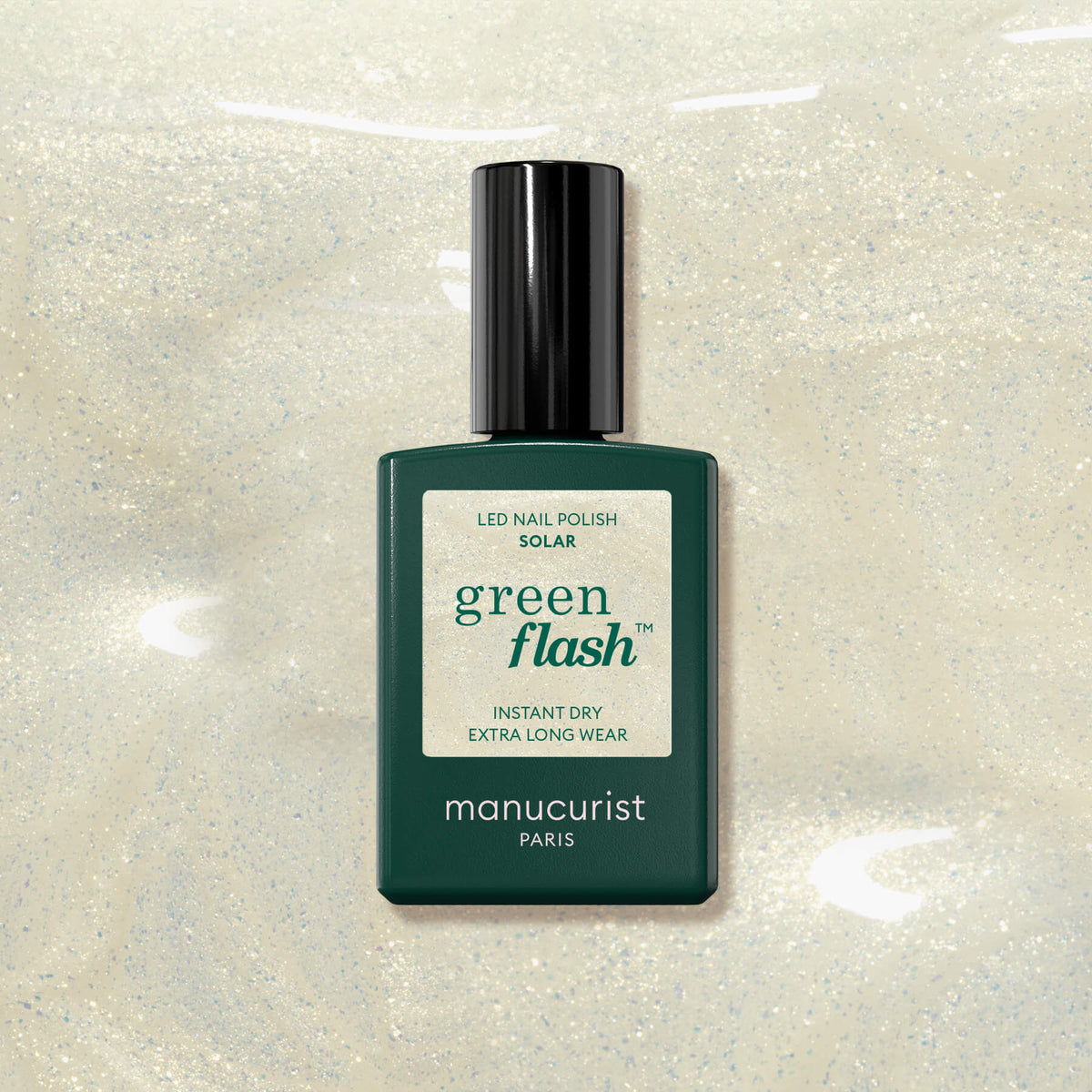 A bottle of Manucurist Solar Green Flash LED nail polish rests on a shimmery light beige surface, its dark green bottle and black cap prominent. The label reads "Green Flash" and "INSTANT DRY EXTRA LONG WEAR," ideal with glitter or golden shimmer.