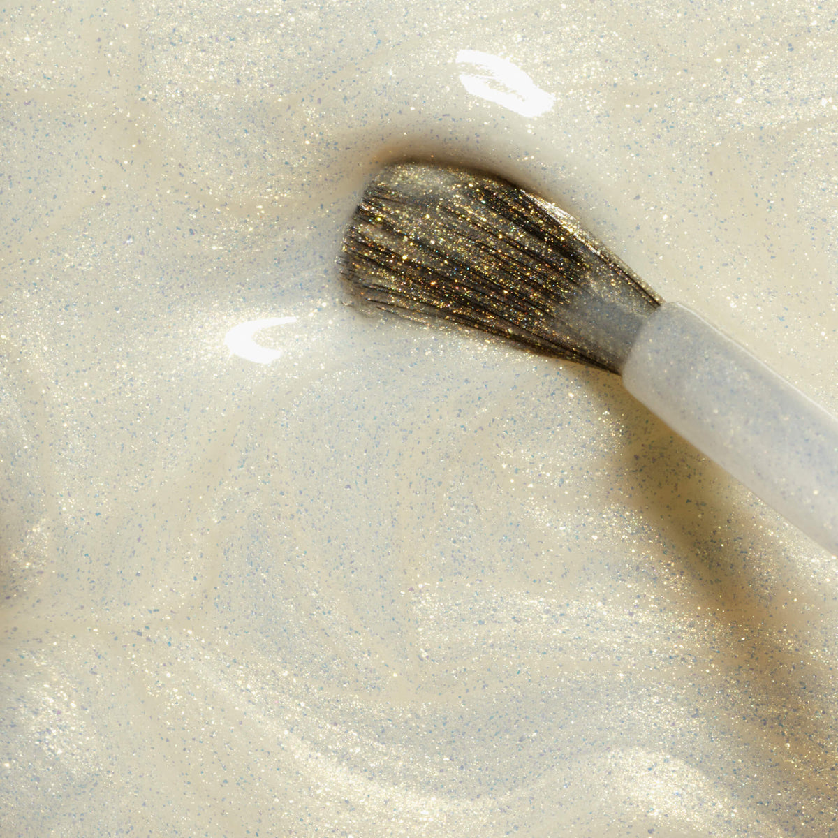 A close-up of the Manucurist Solar brush coated in pearlescent white gel polish with fine glitter, swirling in a pool of the same sheer glittery polish for a dazzling effect.