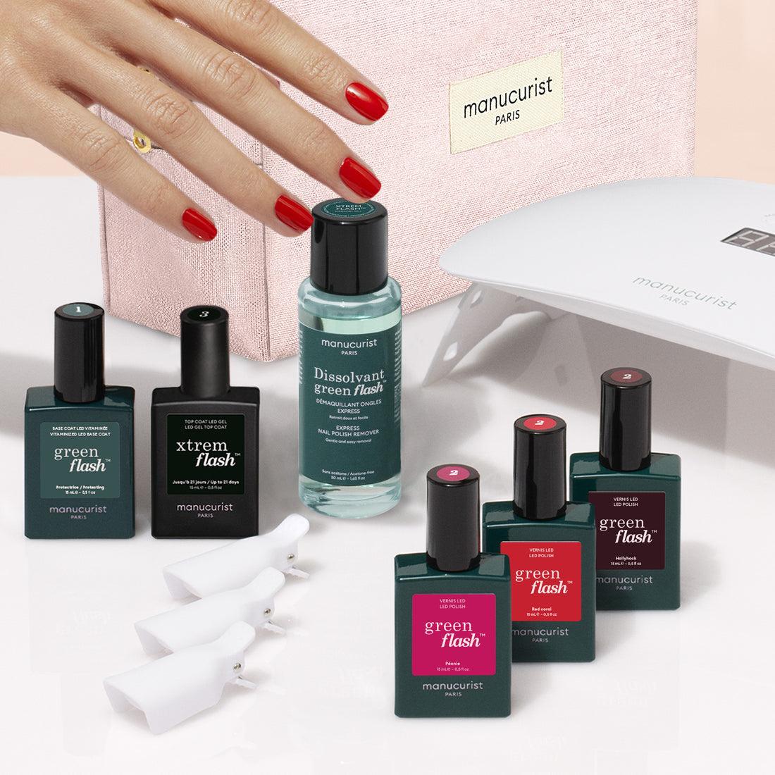 A hand with red manicured nails is shown alongside Manucurist’s Starter Kit, featuring bottles of nail polish, remover, manicure tools, a pink cosmetic pouch, and an LED nail lamp—the ultimate setup for achieving an ultra-glossy manicure or exploring your favorite gel looks.