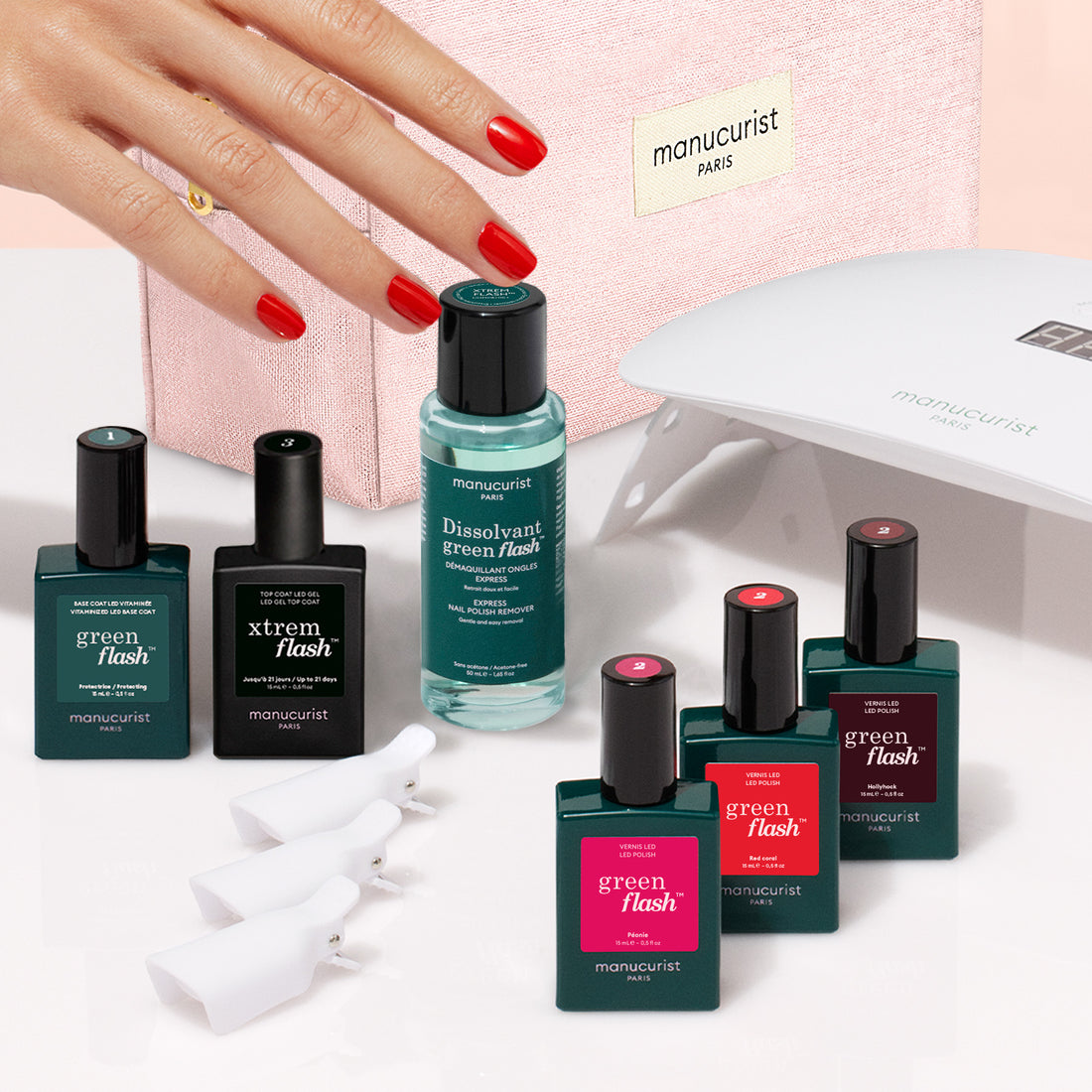 A hand with red manicured nails is shown alongside Manucurist’s Starter Kit, featuring bottles of nail polish, remover, manicure tools, a pink cosmetic pouch, and an LED nail lamp—the ultimate setup for achieving an ultra-glossy manicure or exploring your favorite gel looks.
