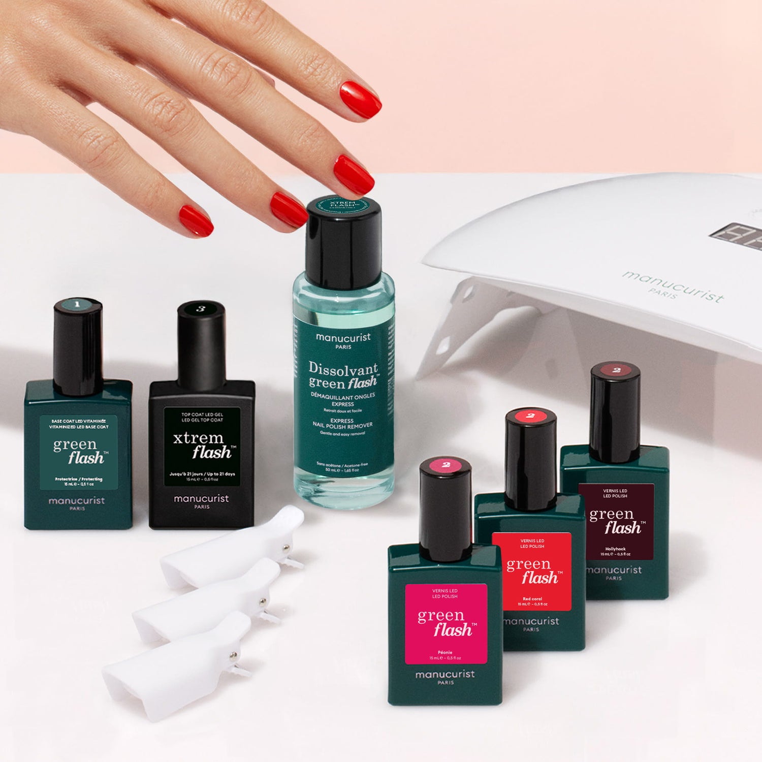 A hand with red nails holds a Manucurist Starter Kit manicure bottle, showcasing the perfect long-lasting manicure. Surrounding it are other green Manucurist Starter Kit gel polish bottles, a dissolvent bottle, white nail clips, and a white UV nail lamp arranged on a beige surface.