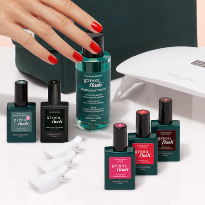 A hand with ultra-glossy red nails reaches for Manucurist’s Starter Kit—featuring polish, remover, clips, and an LED lamp—arranged on a white surface.