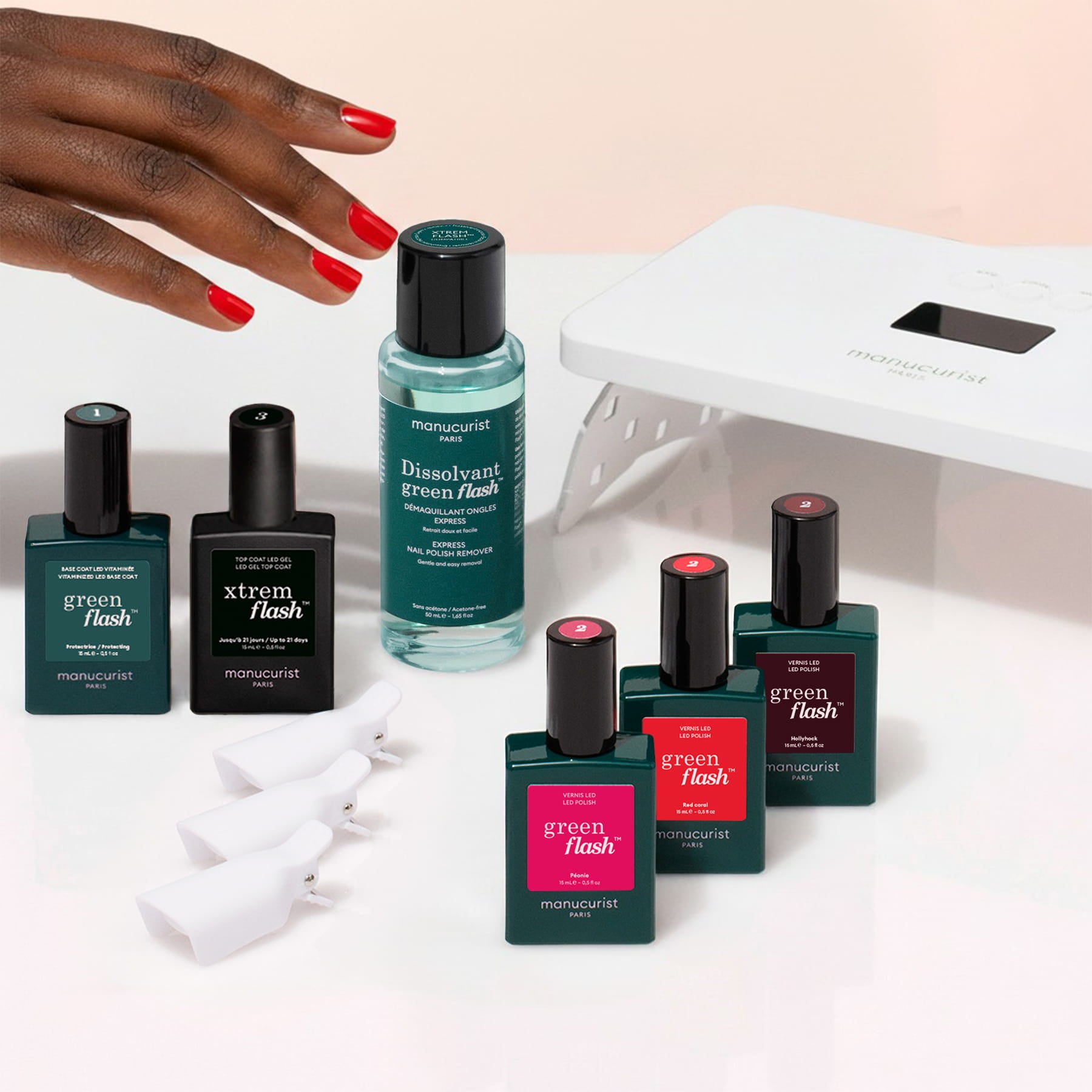 A hand with red nails, accompanied by the Manucurist Starter Kit—which includes bottles of nail polish, a vitaminized base coat, nail polish remover, nail clips, and a UV lamp—displayed on a pale surface: everything you need from Manucurist for a long-lasting manicure.