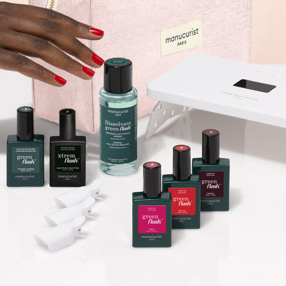 A hand holds a pink Manucurist Starter Kit manicure bag near nail polishes, remover, clips, and an LED lamp from the Manucurist Starter Kit, all displayed on a white surface.